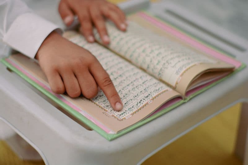 A young boy in a traditional outfit reading the Quran in a classroom setting