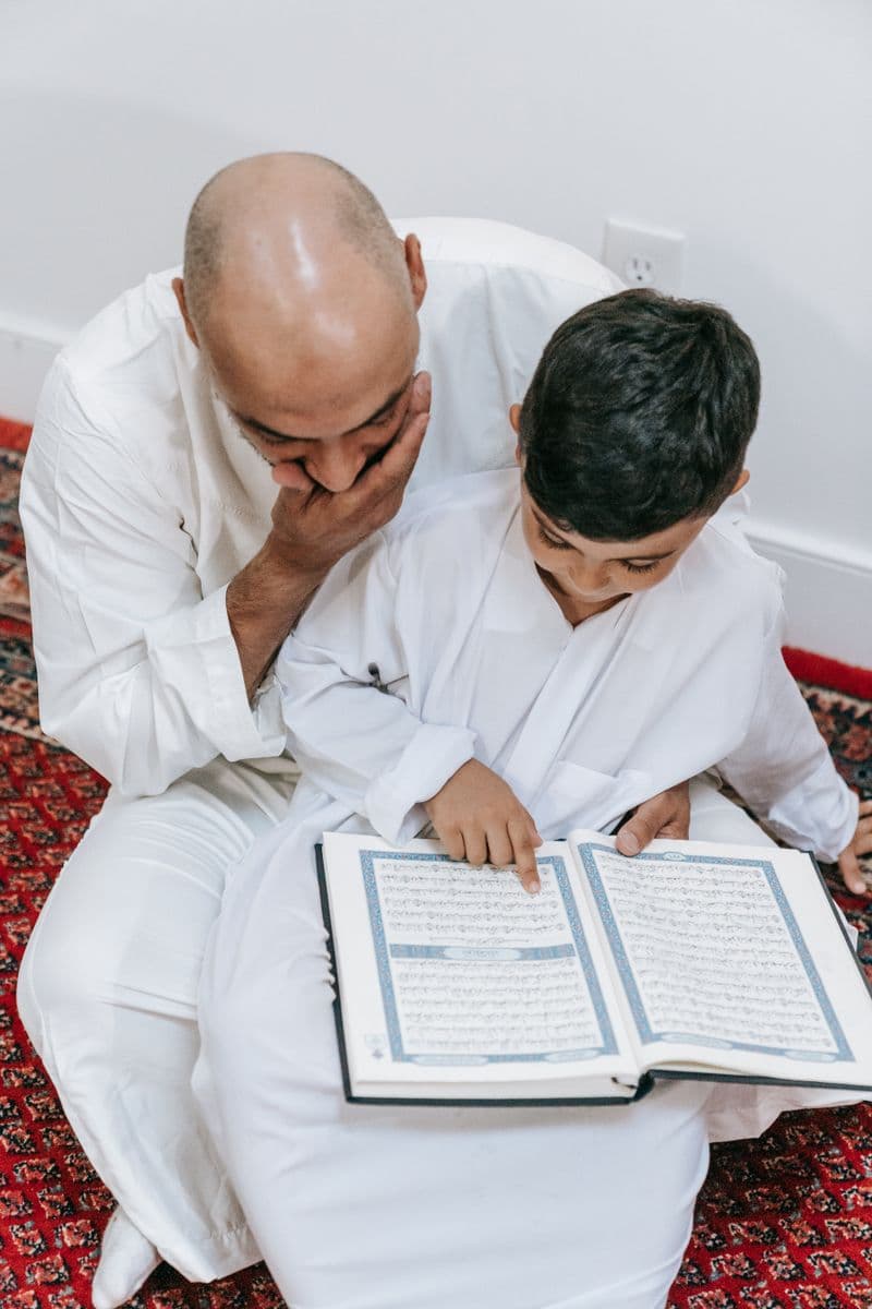 A young boy in traditional attire reading the Quran at a classroom desk